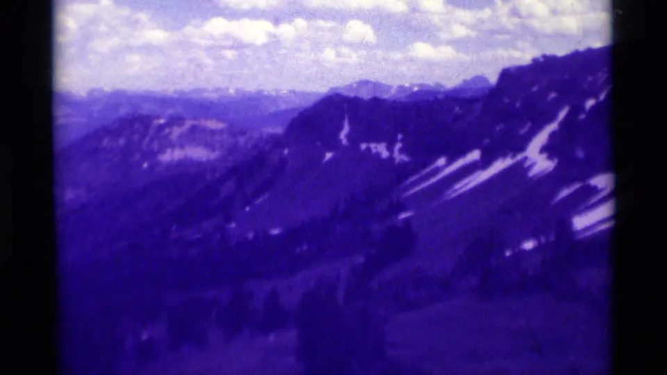 Purple-shifted 1975 8mm frame of Montana mountain ridges with snow traces and a bright clouded sky.