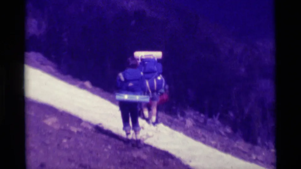 Two hikers with backpacks walk away along a narrow snow-lined trail in the 1975 Agate Springs, Montana home movie.
