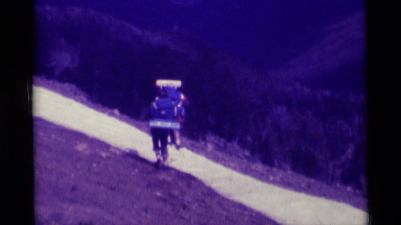 1975 Montana home-movie frame showing a backpacked hiker moving away on a pale trail along a wooded hillside.