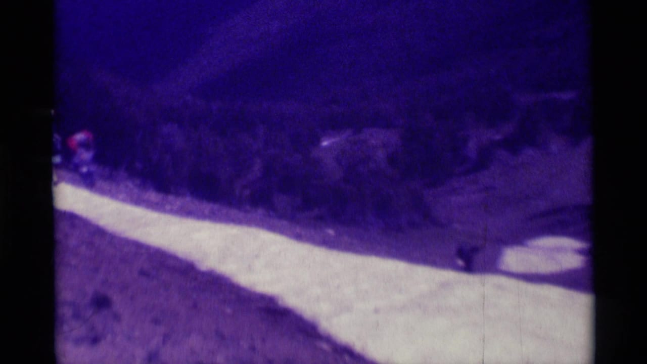 Aged 8mm frame from Montana showing distant hikers along a curving mountainside path with brush and slope beyond.