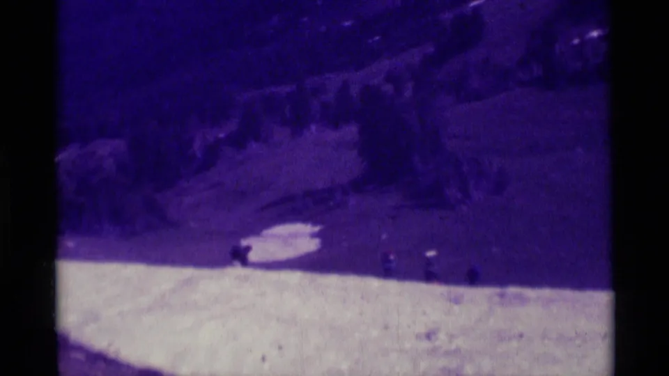 1975 home-movie frame with a bright trail edge, distant backpackers, and a dark wooded hillside in Montana.