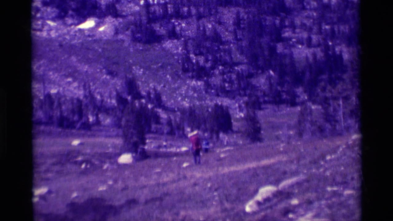 A hiker in a red jacket walks uphill through a rugged Montana trail scene, filmed in soft purple-shifted 8mm color.