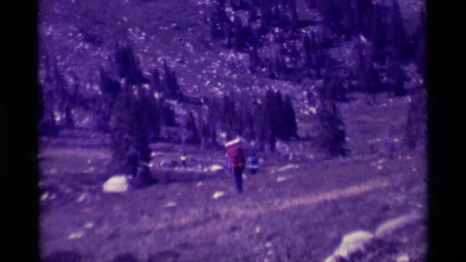Closer 1975 home-movie view of the hiker from behind on a rocky Montana trail with evergreens and pale stones nearby.