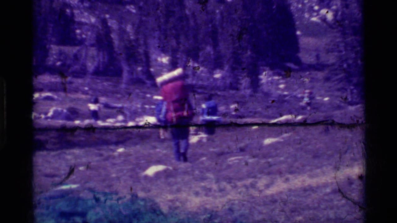A distant hiker in 1975 Agate Springs, Montana home-movie footage walks across rough ground beneath trees and dark mountain terrain.