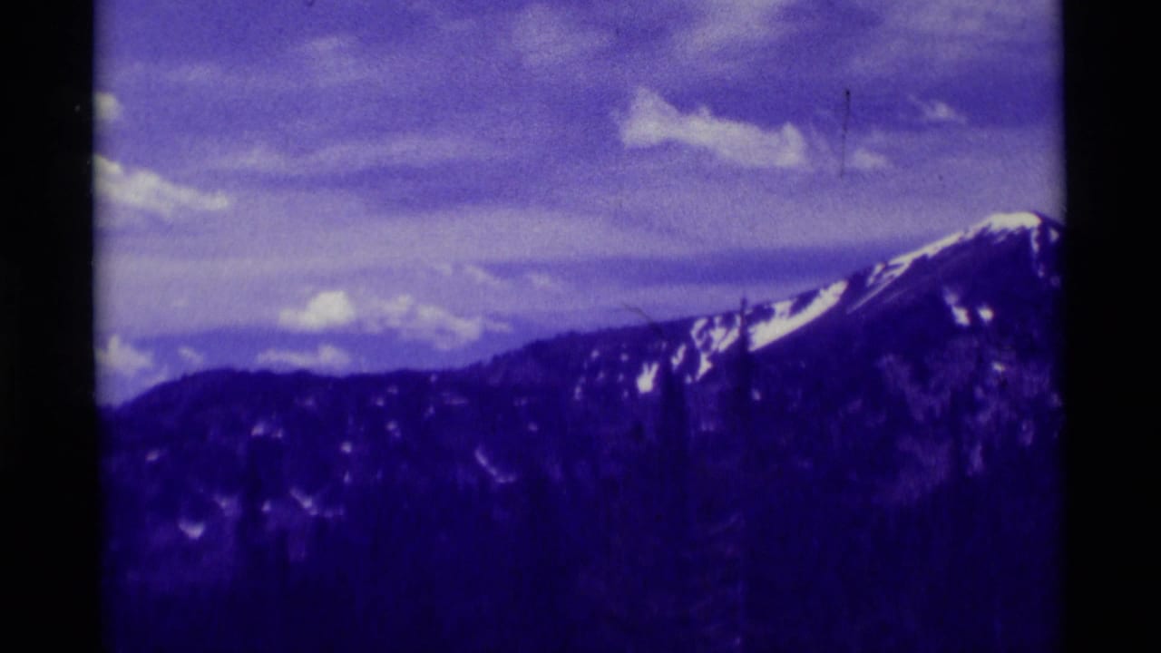 Snow-streaked mountain ridge near Agate Springs, Montana appears under clouds in purple-shifted 1975 8mm home-movie footage.