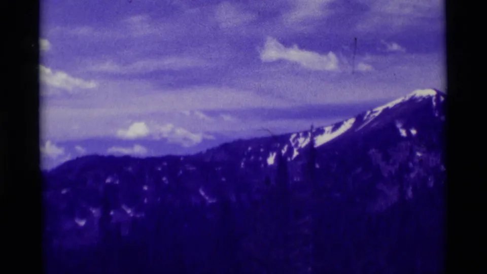 Snow-streaked mountain ridge near Agate Springs, Montana appears under clouds in purple-shifted 1975 8mm home-movie footage.