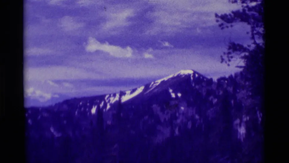 The 1975 Montana home-movie view settles on a distant snow-patched peak with tree branches framing the right side.
