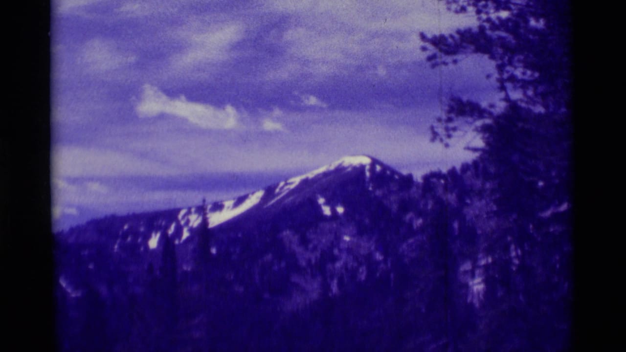 1975 8mm home-movie view near Agate Springs, Montana, showing a snow-streaked mountain peak framed by trees and faded purple color.