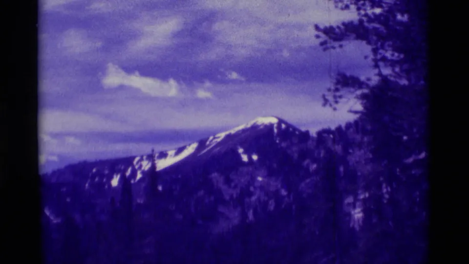 1975 8mm home-movie view near Agate Springs, Montana, showing a snow-streaked mountain peak framed by trees and faded purple color.