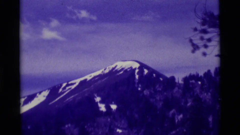 Final 8mm frame from 1975 near Agate Springs, Montana, with a snow-capped peak, cloudy sky, dark treeline, and sparse branches.