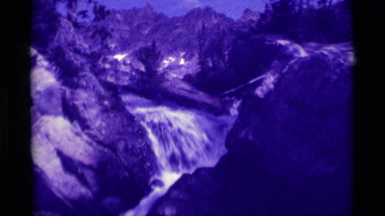 1975 8mm Montana home-movie frame showing a purple-tinted waterfall cascading through rocks below forested mountain terrain.
