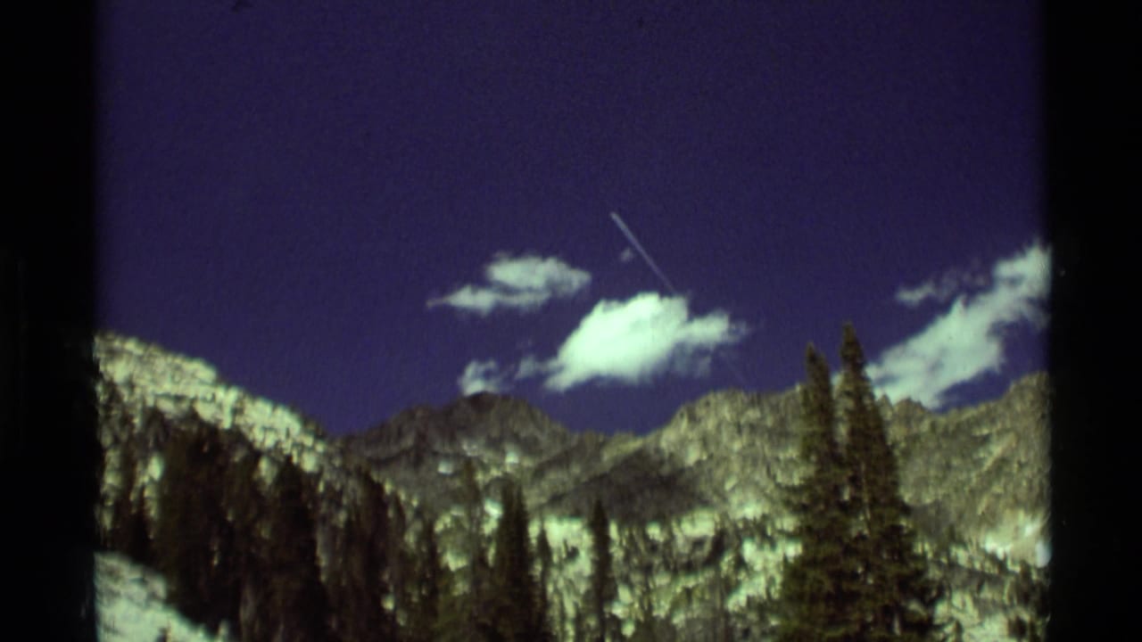 1975 8mm Montana mountain view with a small aircraft or bright contrail crossing above ridges, clouds, and dark evergreen trees.