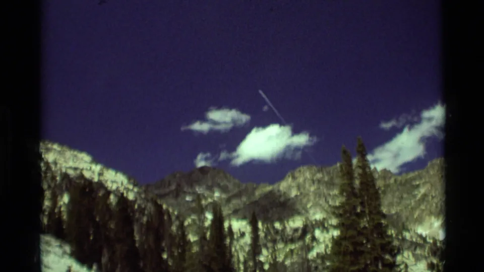 1975 8mm Montana mountain view with a small aircraft or bright contrail crossing above ridges, clouds, and dark evergreen trees.