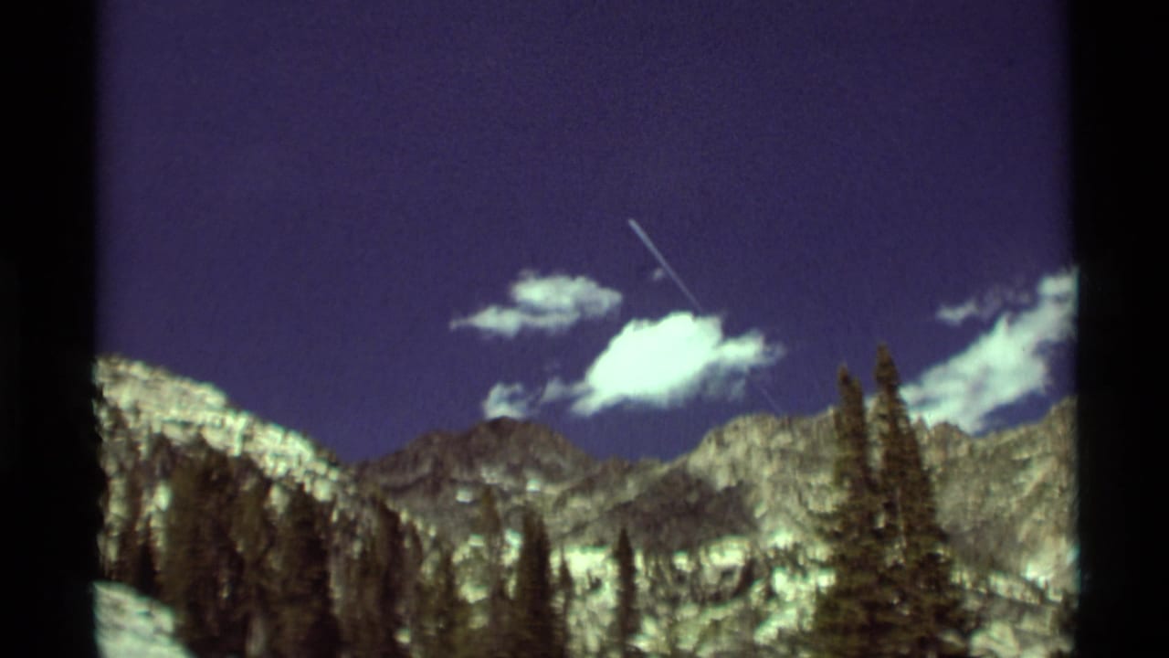 1975 8mm view near Agate Springs, Montana, showing a mountain skyline, conifers, white clouds, and a distant aircraft or contrail.