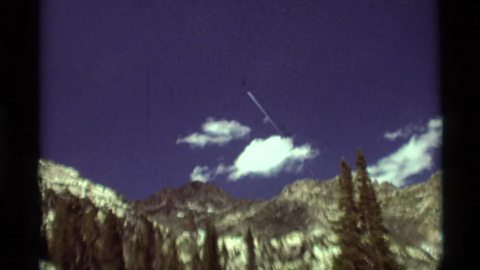 1975 Montana 8mm frame with rugged peaks, dark trees, scattered clouds, and a small aircraft or contrail above the horizon.