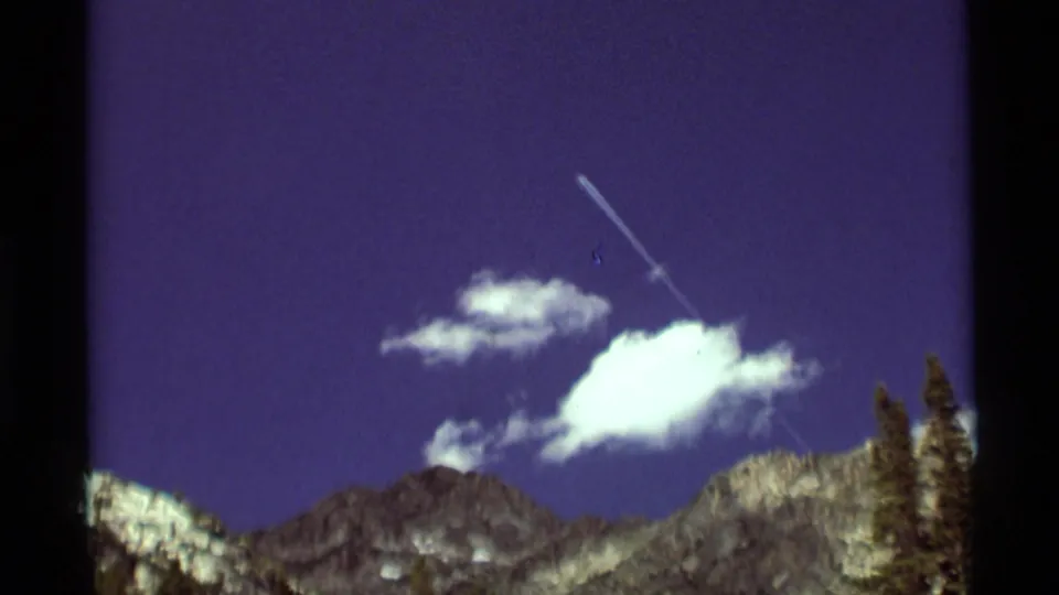 1975 Agate Springs, Montana 8mm frame showing a bright cloud, distant aircraft or contrail, rocky ridges, and conifers along the edge.