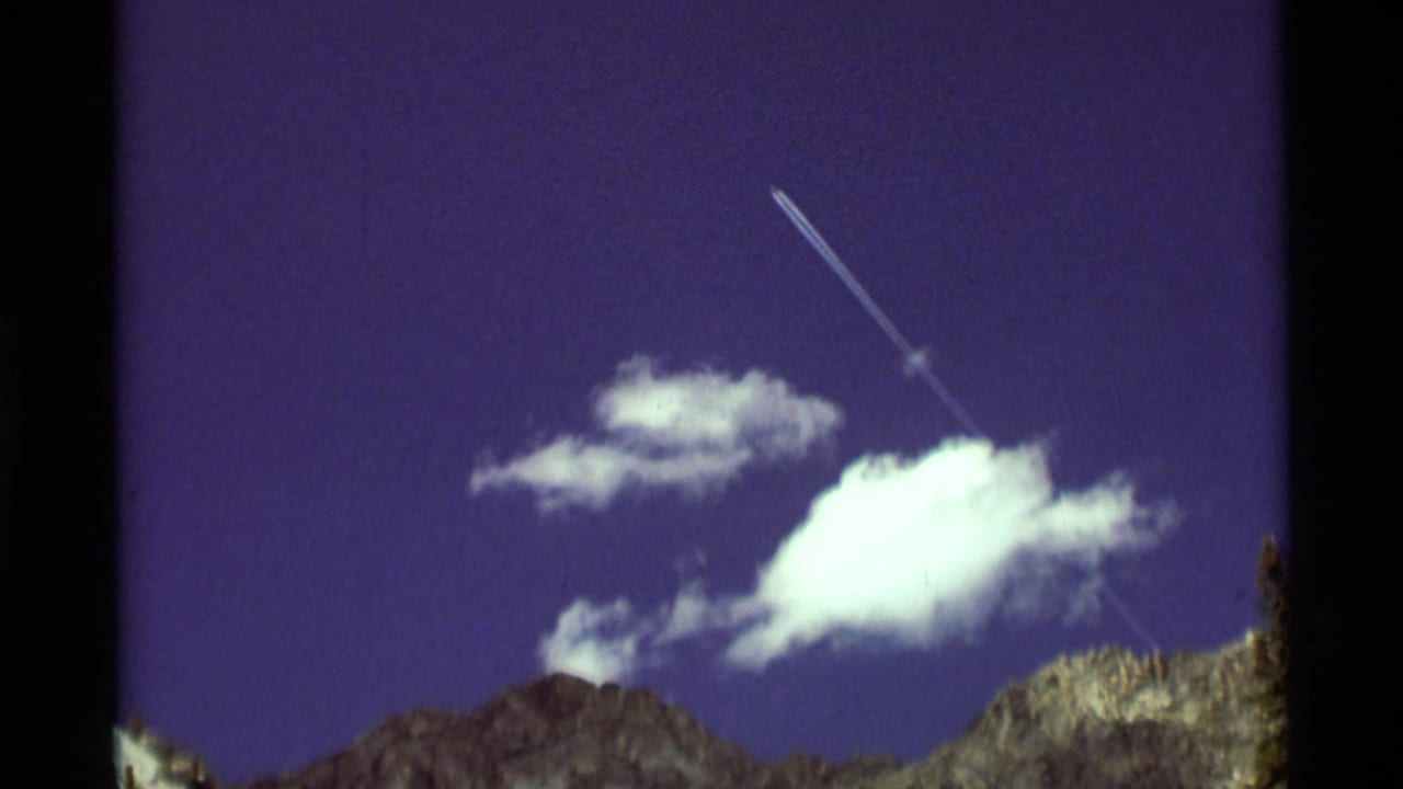 1975 8mm Montana mountain view with rugged peaks, drifting clouds, and a bright diagonal contrail in a deep purple-blue sky.