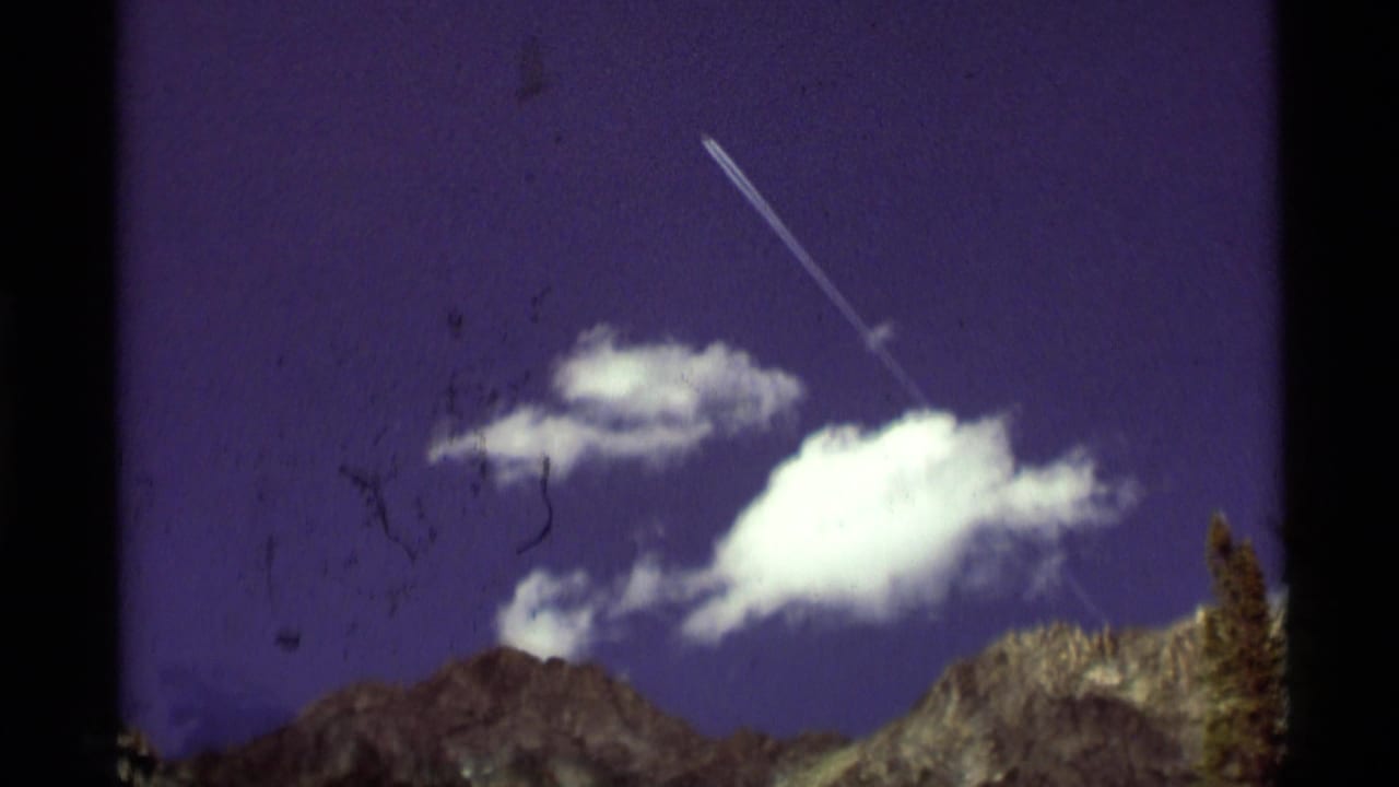 1975 8mm view near Agate Springs, Montana, showing mountain peaks, bright clouds, and a diagonal contrail across a purple-blue sky.