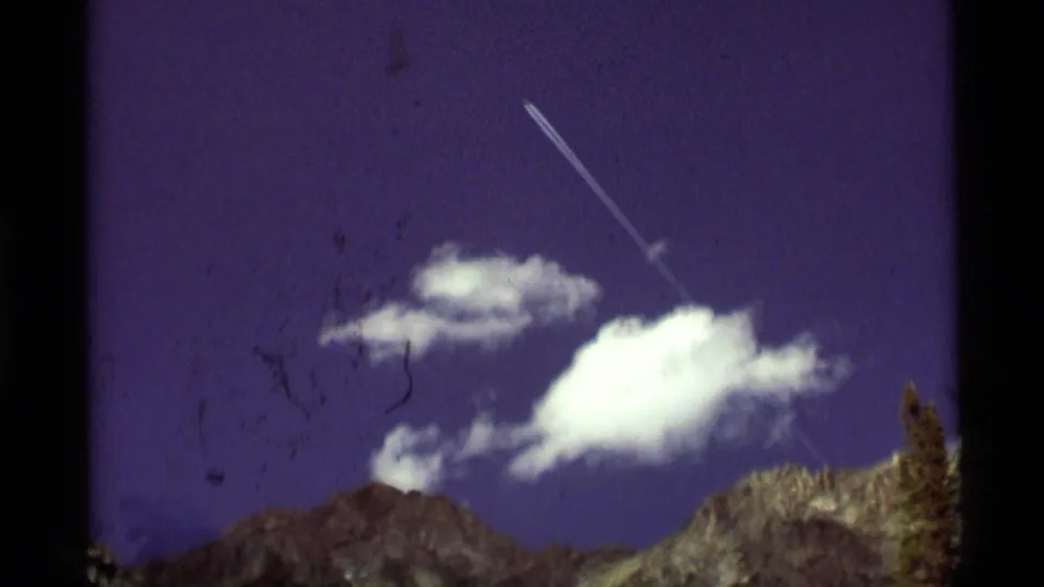 1975 8mm view near Agate Springs, Montana, showing mountain peaks, bright clouds, and a diagonal contrail across a purple-blue sky.