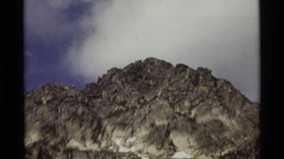 Close 1975 8mm view of a rocky Montana mountain slope with pale cloud cover and a small area of blue sky.