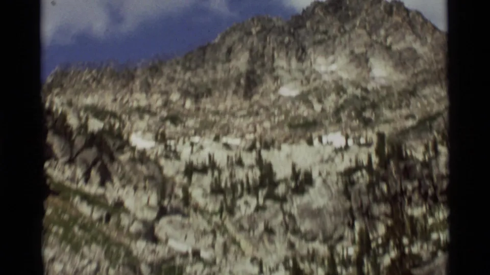 1975 8mm Montana mountain close view with snow patches, sparse dark trees, rocky ridges, and pale sky above.
