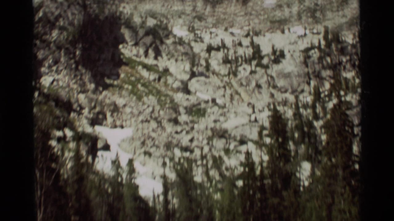 1975 Montana home-movie frame showing a rocky slope with lingering snow and dark evergreen trees in the foreground.