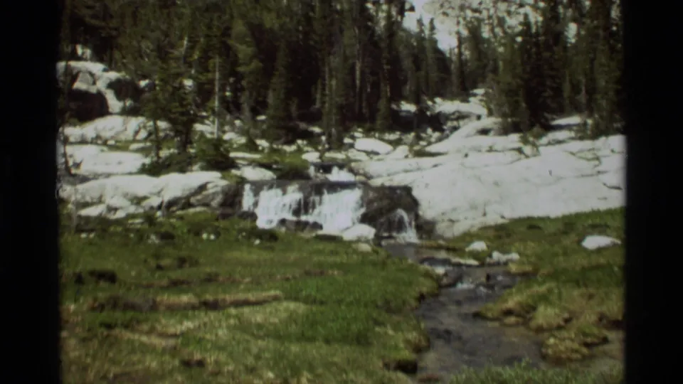 1975 Montana 8mm frame showing a narrow stream crossing a green meadow beneath forest and snow-covered rocks.