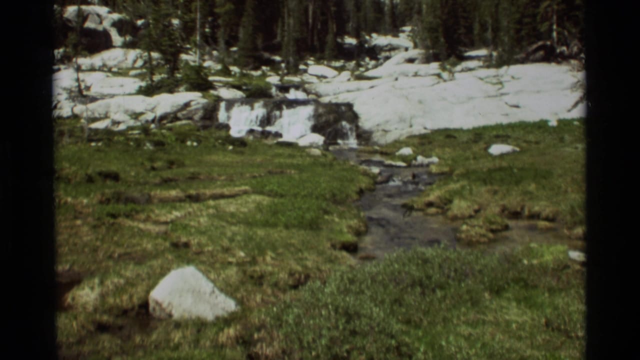 1975 8mm view of a narrow meltwater stream crossing a grassy mountain meadow near Agate Springs, Montana, with snow patches beyond.