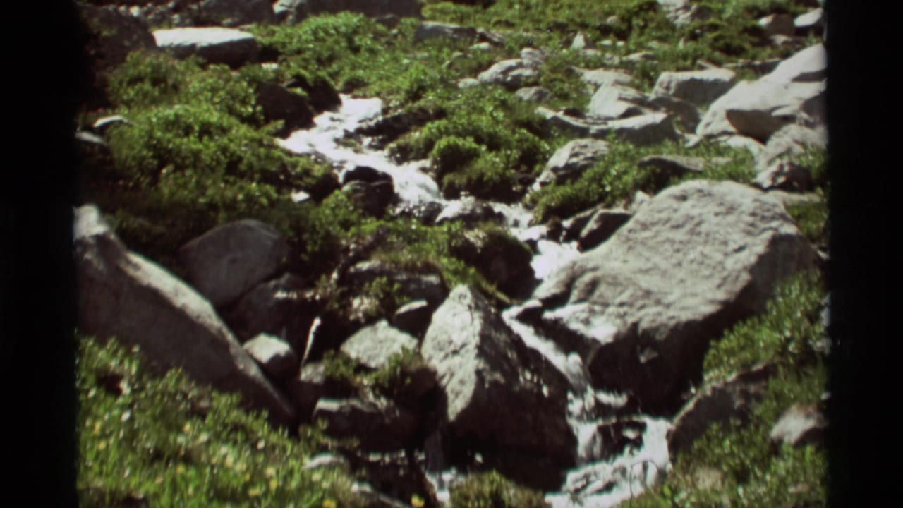 1975 8mm view of a mountain stream in Agate Springs, Montana, flowing around rocks and boulders amid green plants.