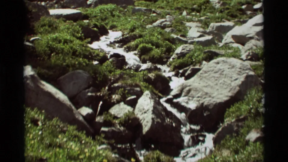 1975 8mm view of a mountain stream in Agate Springs, Montana, flowing around rocks and boulders amid green plants.