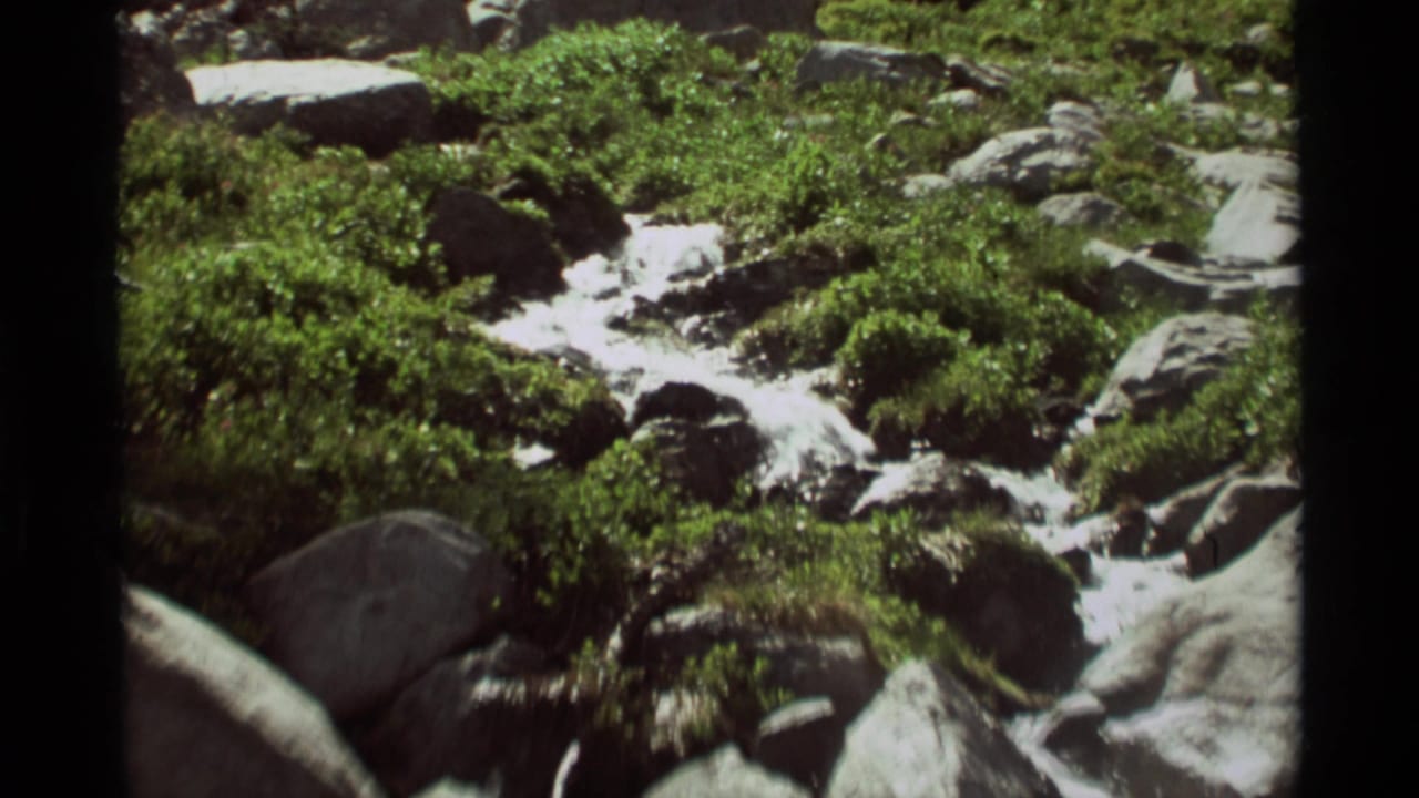 A vintage 8mm frame shows clear water moving over a rocky streambed in Agate Springs, Montana, with dense green vegetation nearby.