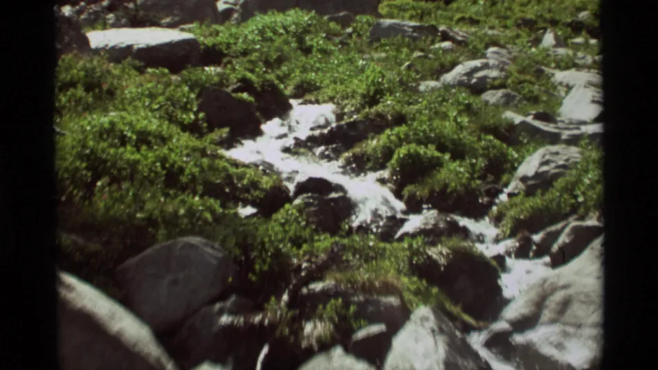 A vintage 8mm frame shows clear water moving over a rocky streambed in Agate Springs, Montana, with dense green vegetation nearby.