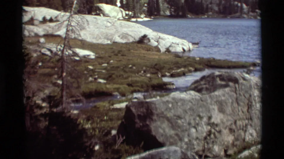 1975 8mm Montana lakeshore view showing granite boulders, grassy ground, sparse trees, and calm blue water.