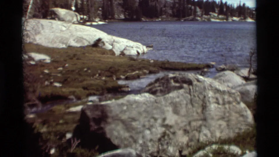 1975 8mm Montana footage of a still lake edged by granite rocks, grassy shoreline, and scattered trees.