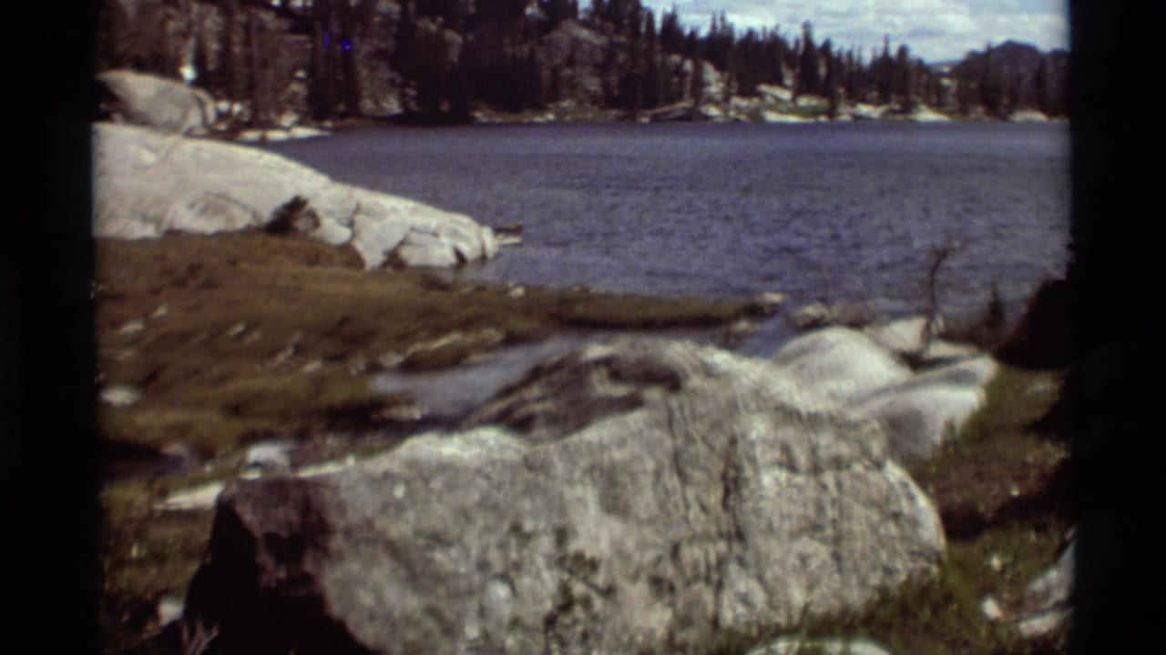 Rocky shoreline beside a calm mountain lake with evergreen trees and distant hills.