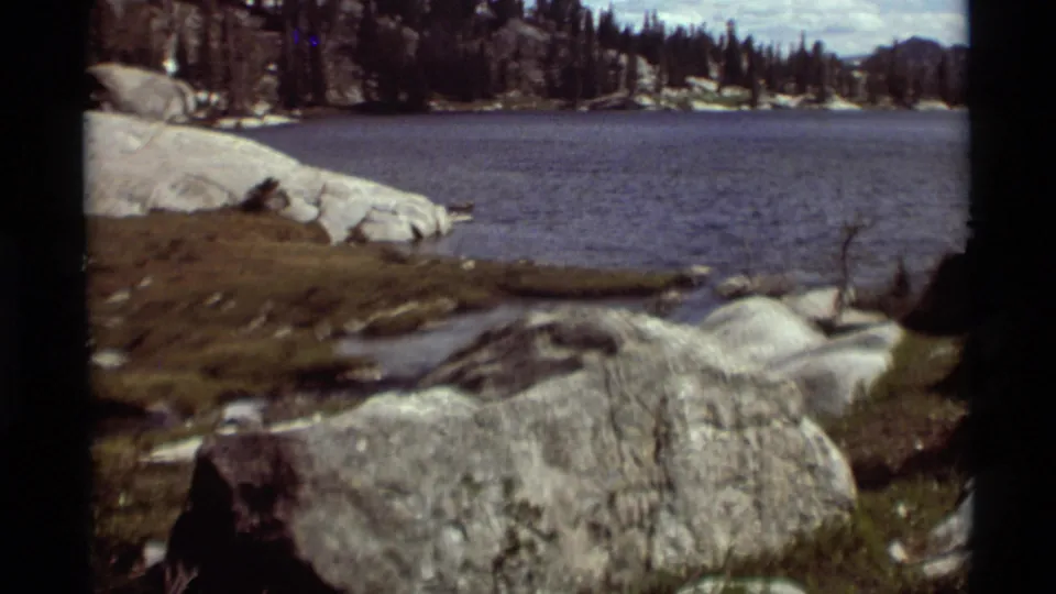 1975 8mm home-movie view of a Montana mountain lake, with pale rocks, shoreline grass, evergreen trees, and distant wooded slopes.