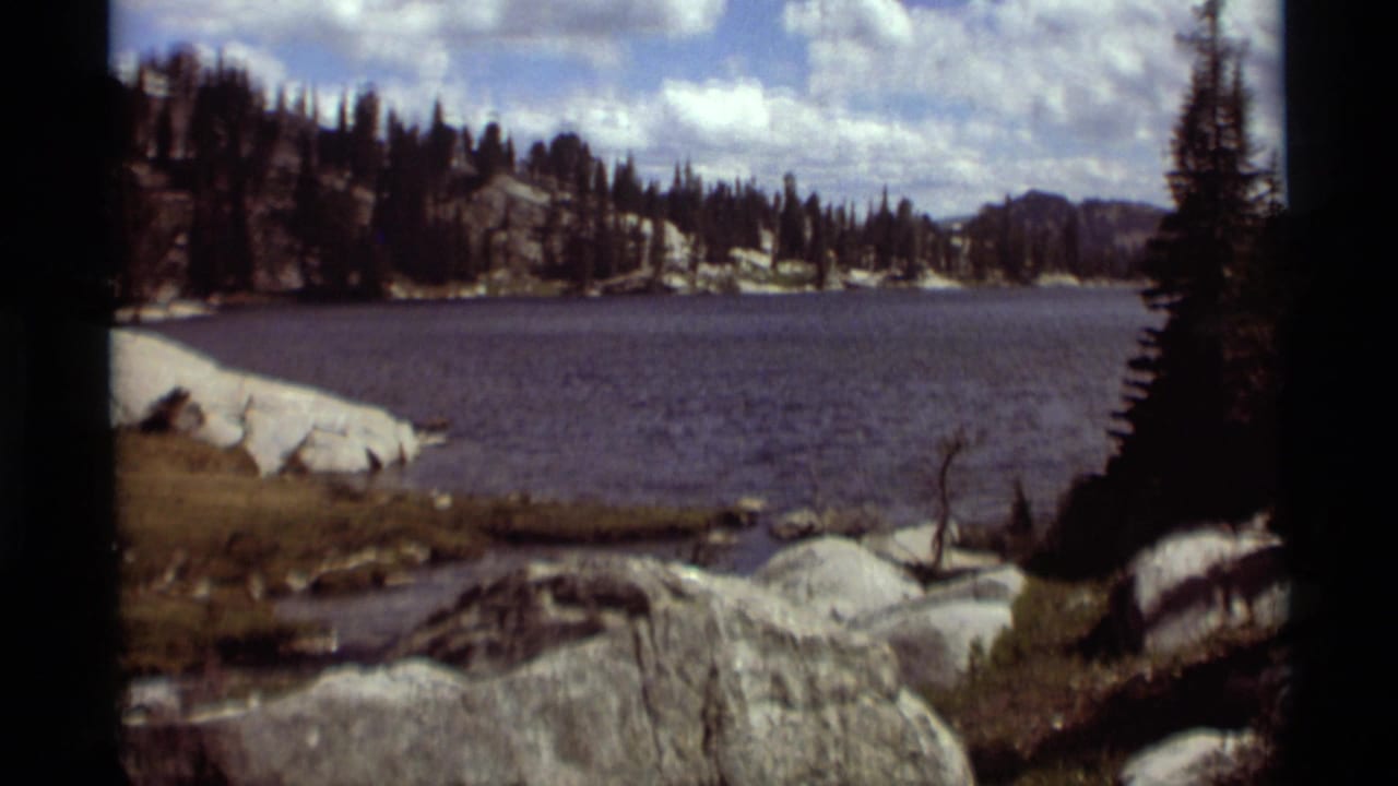 1975 8mm view across an Agate Springs, Montana lake toward rocky banks, evergreen forest, and distant mountain forms beneath broken clouds.