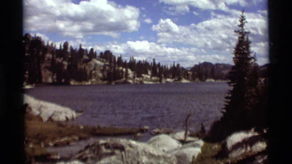 1975 8mm Montana lake scene with open water, pale shoreline boulders, tall conifers, low mountain ridges, and a bright cloud-filled sky.