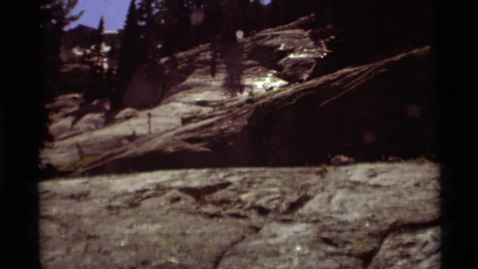 1975 8mm home-movie frame of a sunlit rocky slope near the Montana lake, with scattered evergreens, shadowed rock, and rough shoreline terrain.