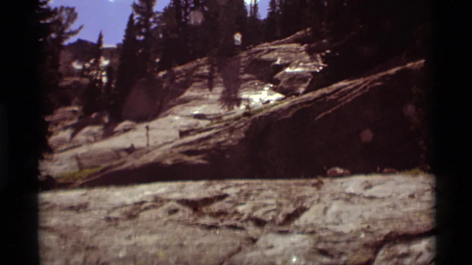1975 8mm view of a rocky forested slope in Agate Springs, Montana, with scattered logs and bright clouds overhead.
