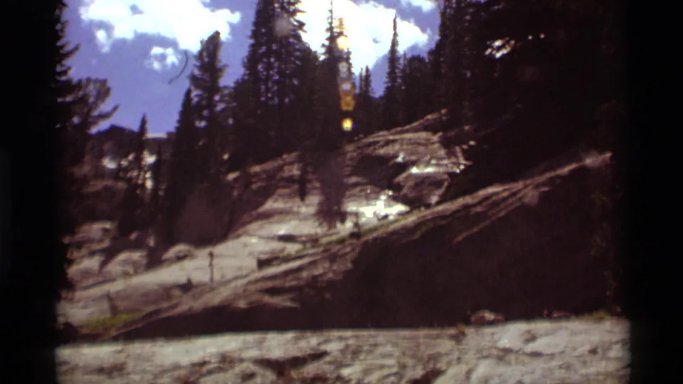 8mm frame of the Agate Springs mountain slope, showing evergreens, exposed rock, scattered logs, and lens flare in the sky.