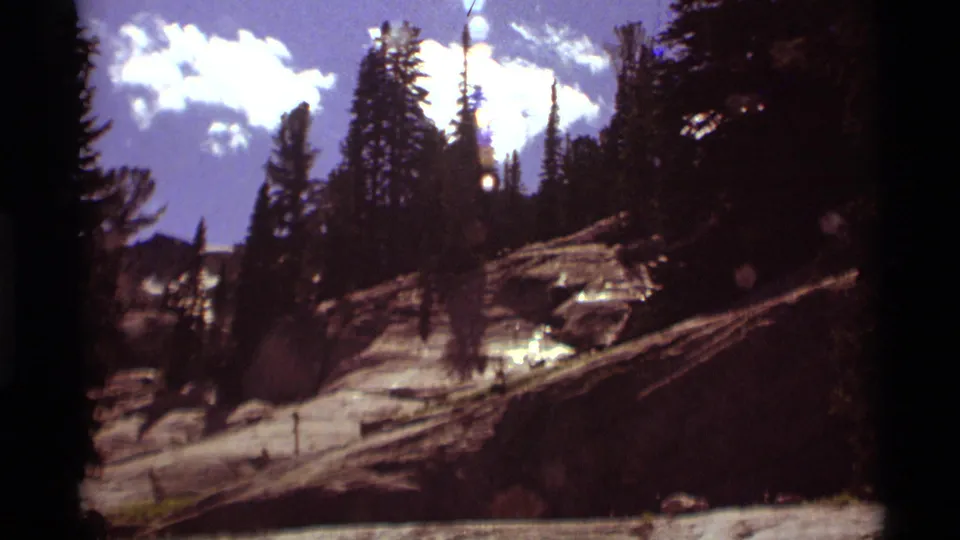 Vintage 8mm mountain view in Montana, with tall evergreens, slanted rock, fallen logs, and bright clouds beyond the ridge.