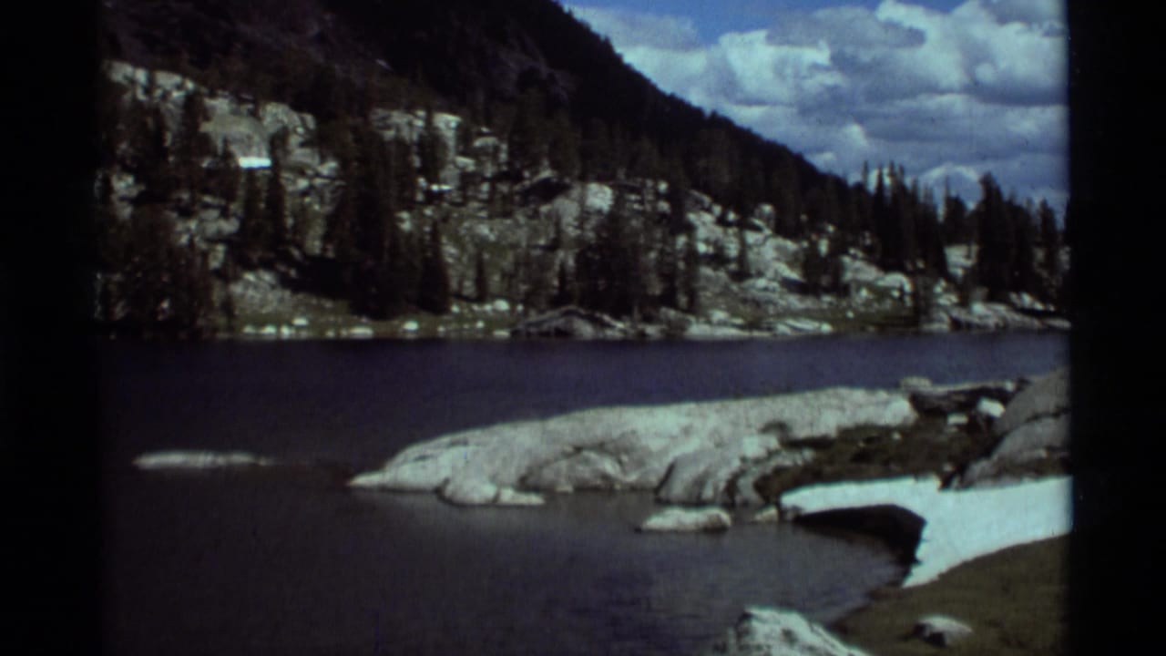 Wide 8mm view of a Montana mountain lake area with rocky shoreline, evergreens, snow patches, and clouds overhead.