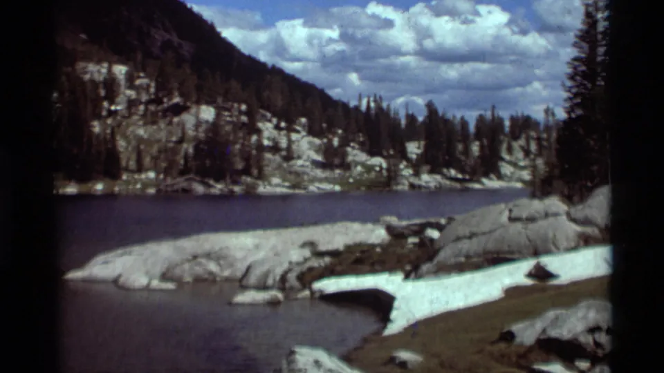 1975 8mm home-movie frame of a mountain lake with pale rocks, lingering snow, evergreen trees, and a partly cloudy sky.