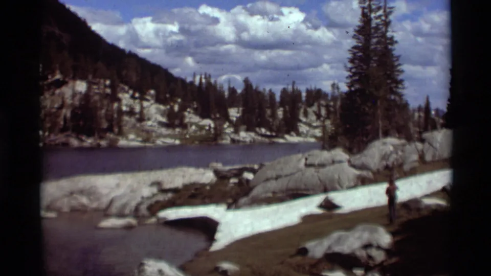 Montana lakeshore scene from 1975 8mm footage, with rocks, snow patches, evergreens, clouds, and one distant figure near the water.