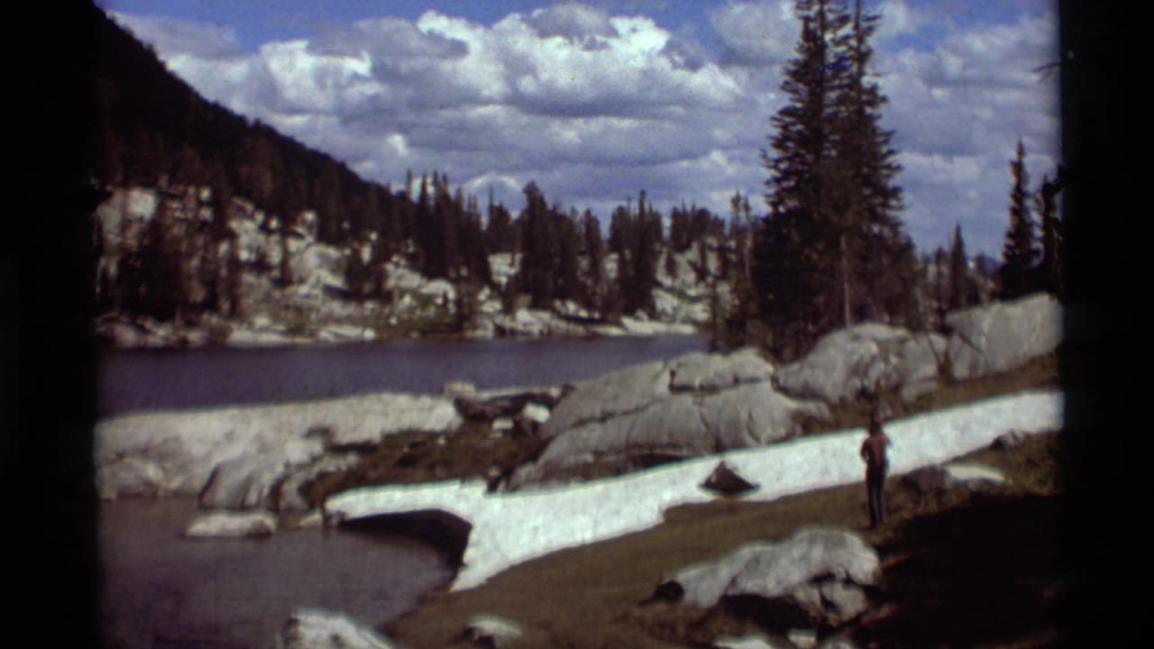1975 Montana 8mm frame showing a lone hiker by alpine water with snow patches, boulders, conifers, and clouds overhead.
