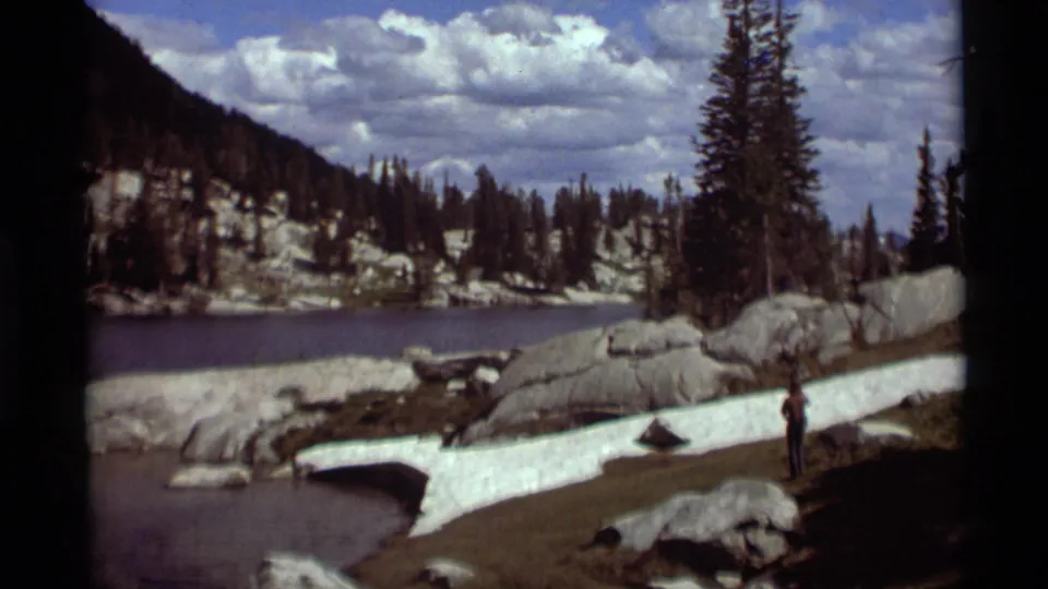 1975 Montana 8mm frame showing a lone hiker by alpine water with snow patches, boulders, conifers, and clouds overhead.