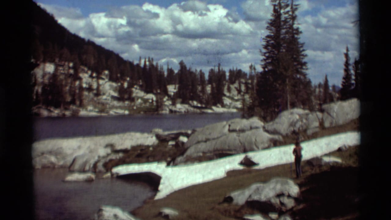 Mountain lake or river edge with snow, boulders, evergreens, and one person standing near the water.