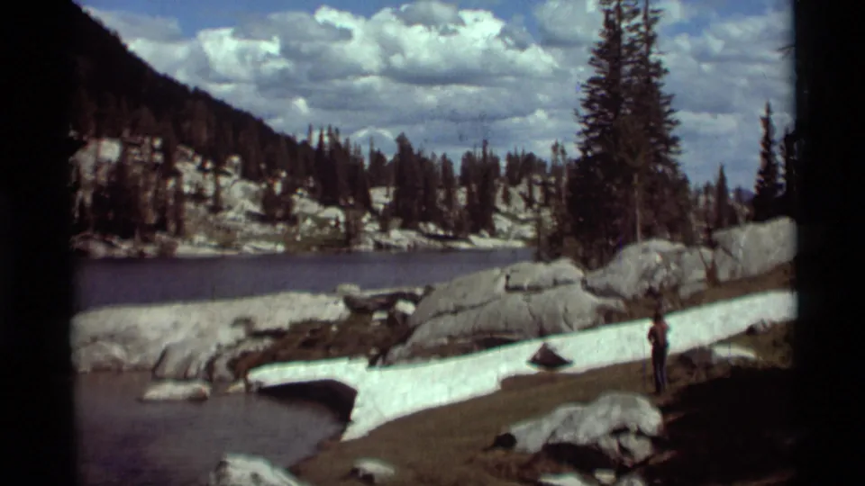 1975 Montana home-movie view of a hiker beside alpine water, with snow-dusted rocks, pine forest, and partly cloudy sky.