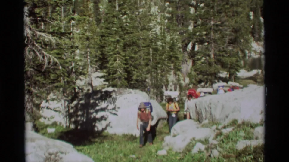 1975 Montana 8mm frame of backpackers moving along a rocky forest trail with boulders, conifers, grass, and lingering snow.